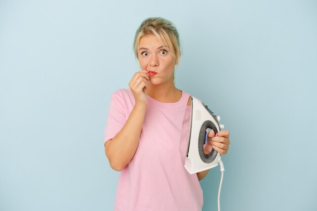 Young Russian woman holding iron isolated on blue background with fingers on lips keeping a secret.