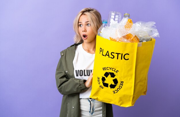 Young Russian woman holding a recycling bag full of paper to recycle isolated on purple doing surprise gesture while looking to the side