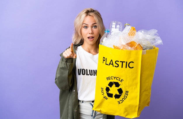 紫色の壁に隔離されたリサイクルする紙でいっぱいのリサイクルバッグを持っている若いロシアの女性は驚いて正面を指