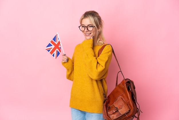 Молодая русская женщина держит флаг Соединенного Королевства, изолированную на розовой стене, смотрит в сторону и улыбается