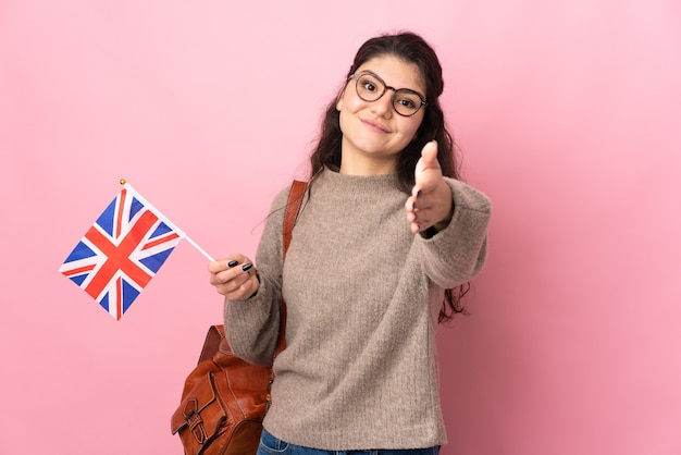 Молодая россиянка с флагом Соединенного Королевства изолирована на розовой стене и пожимает руку для заключения хорошей сделки