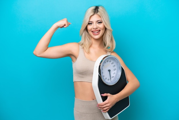 Young Russian woman isolated on blue background holding a weighing machine and doing strong gesture