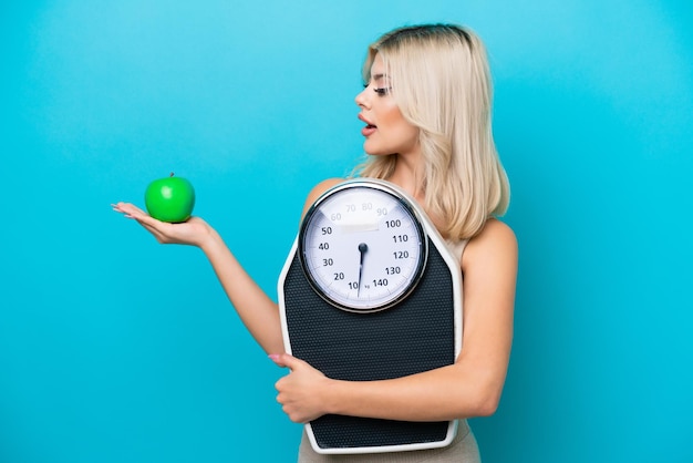 Young Russian woman isolated on blue background holding a weighing machine while looking an apple