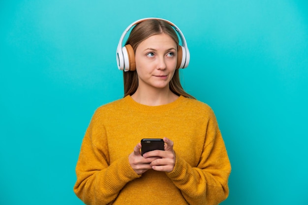 Young Russian woman isolated on blue background listening music with a mobile and thinking