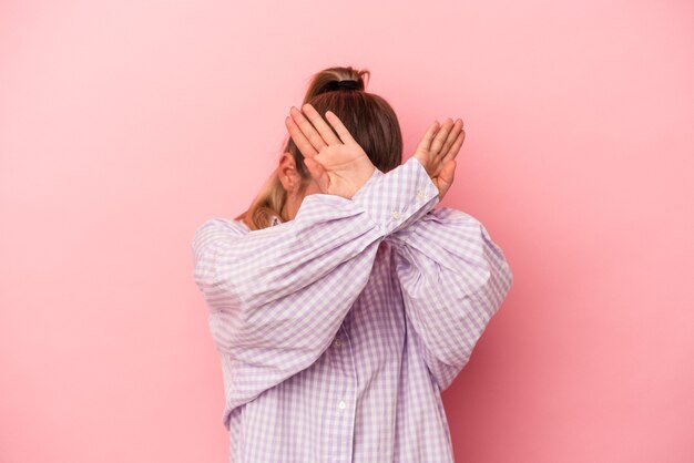 Young Russian woman isolated on pink background covering ears with hands trying not to hear too loud sound.