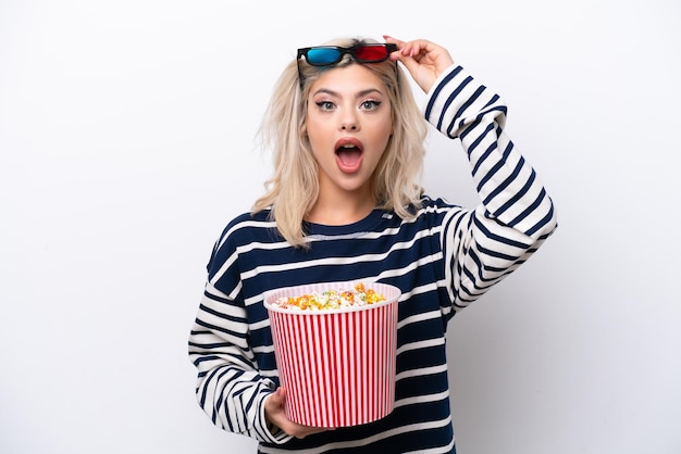 Young Russian woman isolated on white background surprised with 3d glasses and holding a big bucket of popcorns