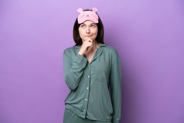 Young Russian woman in pajamas isolated on purple background having doubts and thinking