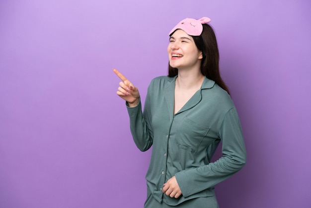 Young russian woman in pajamas isolated on purple background pointing finger to the side and presenting a product