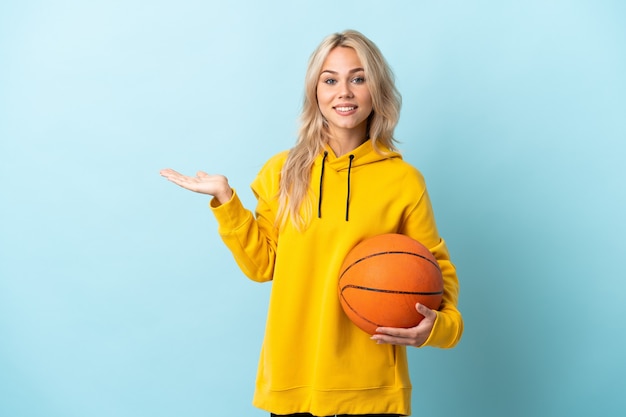 Young Russian woman playing basketball isolated on blue background holding copyspace imaginary on the palm to insert an ad