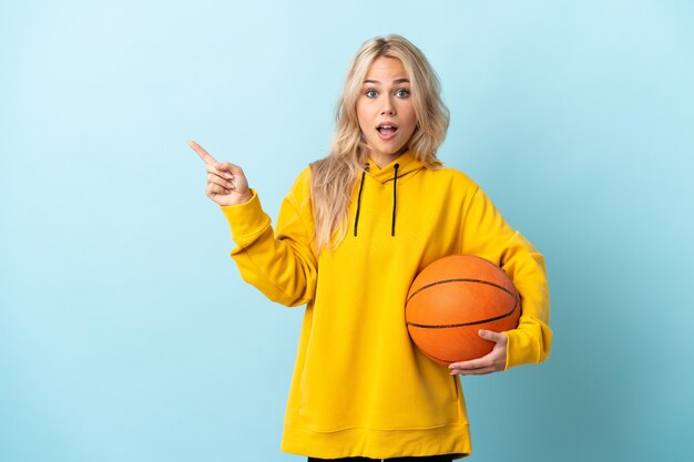 Young Russian woman playing basketball isolated on blue surprised and pointing finger to the side