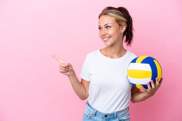 Young Russian woman playing volleyball isolated on pink background pointing finger to the side and presenting a product