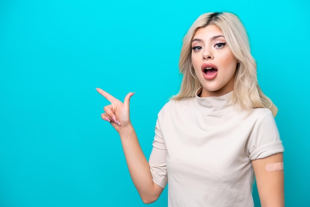 Young Russian woman wearing band aid isolated on blue background surprised and pointing finger to the side