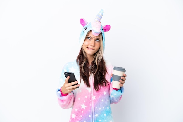 Young Russian woman wearing a unicorn pajama isolated on white background holding coffee to take away and a mobile while thinking something