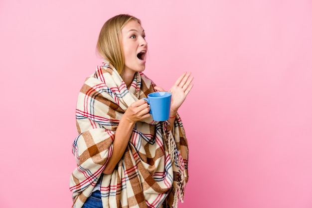 Young russian woman wrapped in a blanket drinking coffee shouts loud, keeps eyes opened and hands tense.