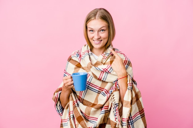 Young russian woman wrapped in a blanket drinking coffee smiles, pointing fingers at mouth