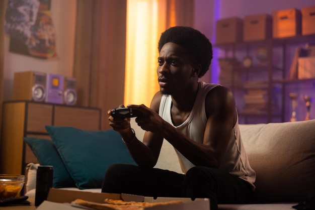 Young sad afro guy relaxing at home sitting on couch and\
playing video games in the evening alone st