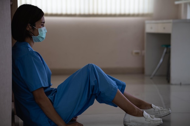 Молодая грустная азиатская медсестра сидит на больничном полу, делая перерыв после тяжелой работы в больнице.