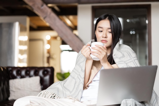 Young sad asian woman drinking tea being sick at home looking\
at laptop screen selfquarantine during...