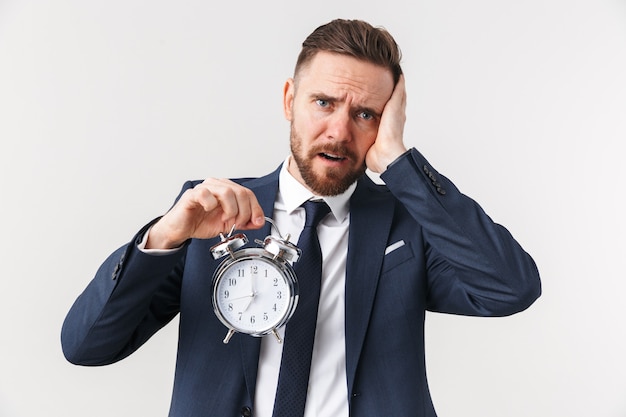 young sad businessman posing isolated over white wall holding alarm clock