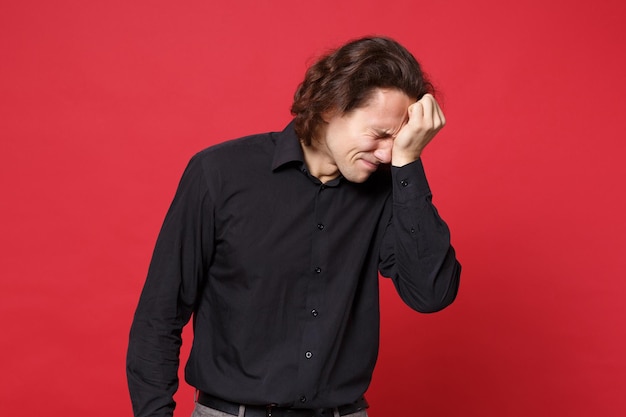 Young sad curly long haired man in black shirt standing posing isolated on red wall background studio portrait. people sincere emotions lifestyle concept. mock up copy space. problem crisis depression