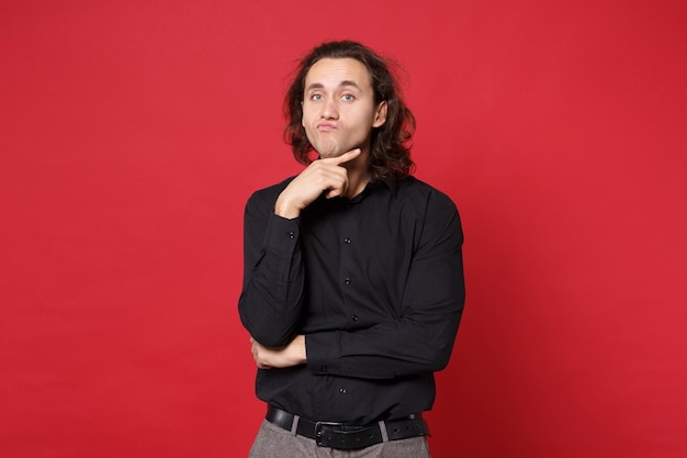 Young sad curly long haired man in black shirt standing posing isolated on red wall background studio portrait. People sincere emotions lifestyle concept. Mock up copy space. Problem crisis depression