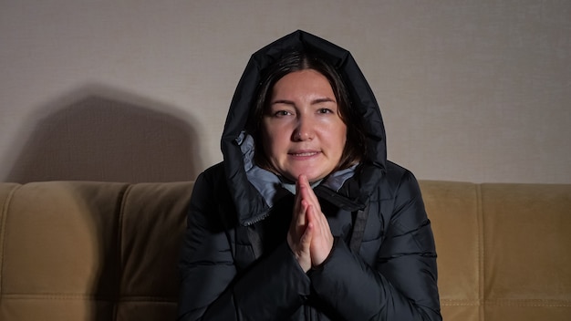 フード付きの黒い暖かいジャケットを着ている若い悲しい凍った女性は、セントラルヒーティングなしで寒い部屋の柔らかいソファに座っている手を加熱しようとします。