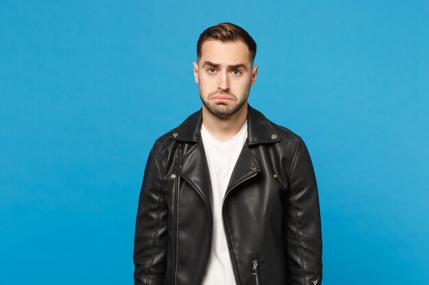 Photo young sad frustrated worried unshaven man in black jacket white t-shirt looking camera isolated on blue wall background studio portrait. people sincere emotions lifestyle concept. mock up copy space.