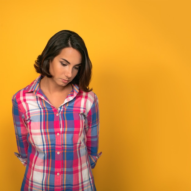 Young sad pensive woman feels guilty, thinks looking down while standing against of yellow wall in background with copy space on right side.