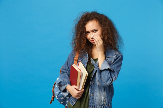 Молодой грустный студент в джинсовой одежде и рюкзаке держит книги, изолированные на синей стене