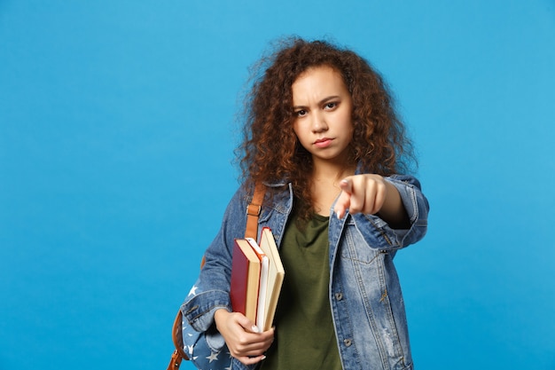 デニムの服とバックパックの若い悲しい学生は青い壁に隔離された本を保持します