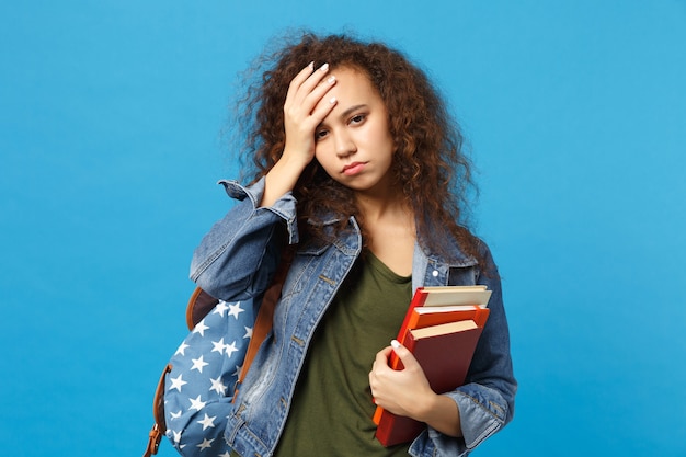 Young sad student in denim clothes and backpack holds books isolated on blue wall