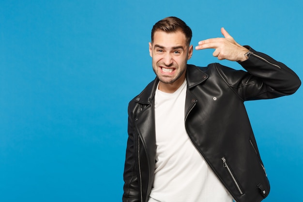 Young sad unshaven man in black jacket white t-shirt looking camera, point fingers to head as if he about shoot himself isolated on blue wall background. People lifestyle concept. Mock up copy space.