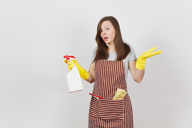 Young sad upset bewildered housewife in yellow gloves striped apron cleaning rag squeegee in pocket isolated on white background. Woman spread hands, hold spray bottle with cleaner liquid. Copy space.