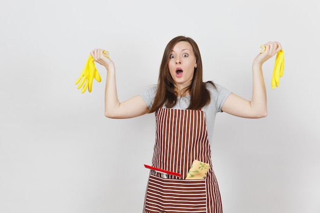 Young sad upset tired shocked housewife in striped apron with cleaning rag in pocket isolated. Pretty housekeeper woman holding yellow gloves in spreading hands