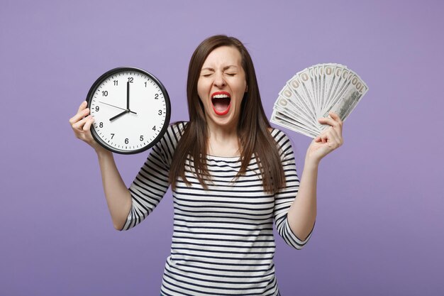 Young sad upset woman girl in casual striped clothes posing isolated on violet purple background studio portrait. People sincere emotions lifestyle concept. Mock up copy space. Hold clock, cash money.