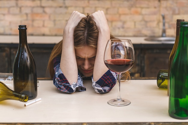 Young sad and wasted alcoholic woman sitting at kitchen couch drinking red wine, completely drunk looking depressed lonely and suffering hangover in alcoholism and alcohol abuse.