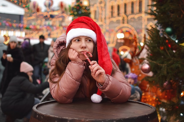 A young sad woman outdoors in a Santa hat puts a sweet cane to her lips and looks bored to the side