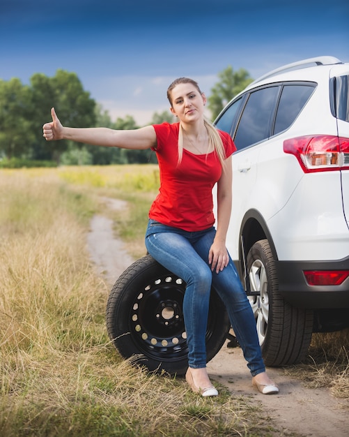 Young sad woman sitting on wheel at broken car and hitchhiking