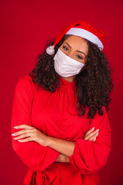covid-19 코로나바이러스 바이러스를 입은 크리스마스 모자를 쓴 젊은 산타클로스 여성은 빨간색 배경 스튜디오에 격리된 마스크를 저장했습니다. 새 해 복 많이 받으세요 축 하 휴일 개념입니다.