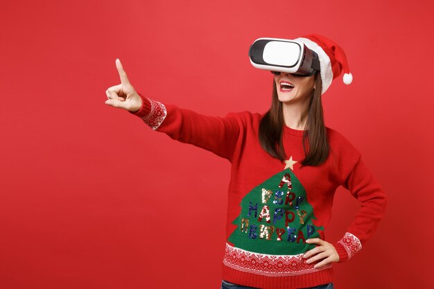 Young Santa girl looking in headset touch something like push click on button, pointing at floating virtual screen isolated on red background. Happy New Year 2019 celebration holiday party concept.