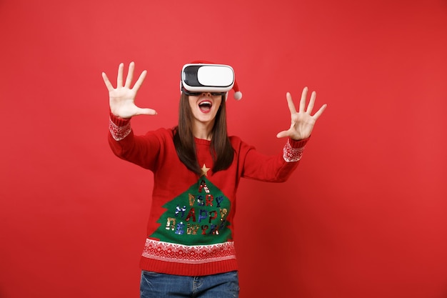 Young Santa girl looking in headset touch something like push click on button, pointing at floating virtual screen isolated on red background. Happy New Year 2019 celebration holiday party concept.