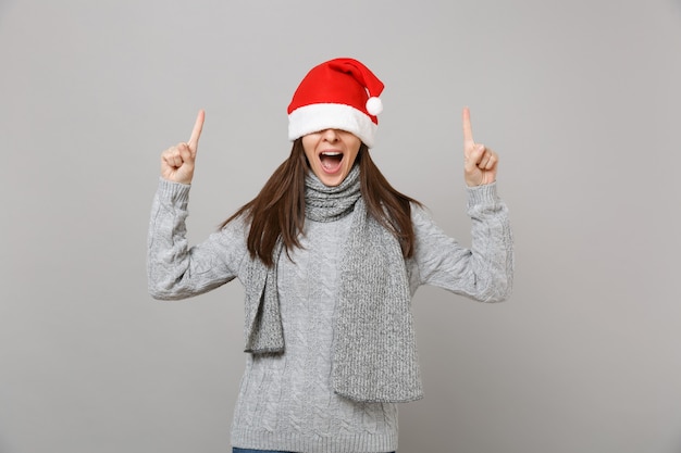 Young Santa girl in sweater scarf covering eyes with Christmas hat pointing index fingers up keeping mouth wide open isolated on grey background. Happy New Year 2019 celebration holiday party concept.