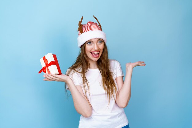 Young santa woman with horns surprised with happiness and surprise with a gift in her hand on a blue...