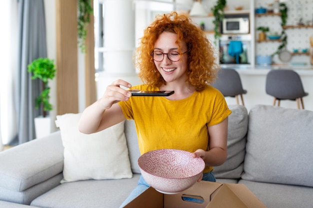 Young satisfied happy woman shopaholic customer sit on sofa unpack parcel delivery box online shopping shipment concept taking photos of product to post on social media