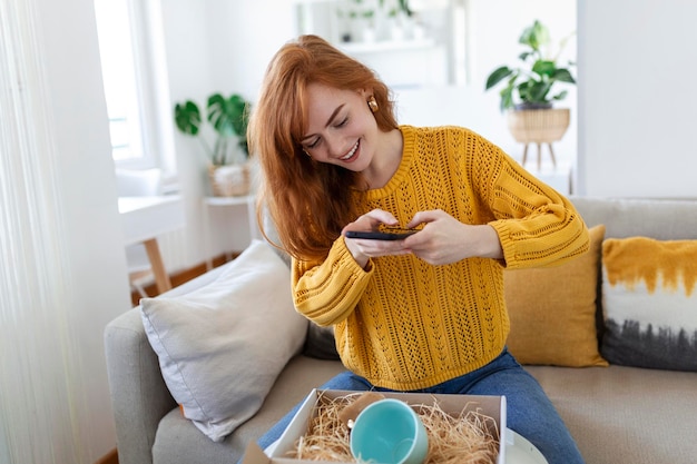 Young satisfied happy woman shopaholic customer sit on sofa unpack parcel delivery box online shopping shipment concept taking photos of product to post on social media