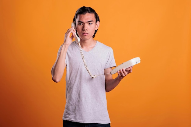 Young scared man talking on landline phone. Confused asian teen chatting, holding retro telephone, frightened person with terrified facial expression answering call on orange background