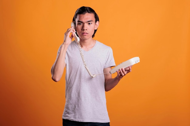 Young scared man talking on landline phone. confused asian teen chatting, holding retro telephone, frightened person with terrified facial expression answering call on orange background