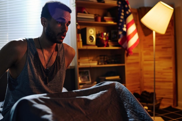 Молодой испуганный мужчина с посттравматическим синдромом сидит на кровати после кошмара