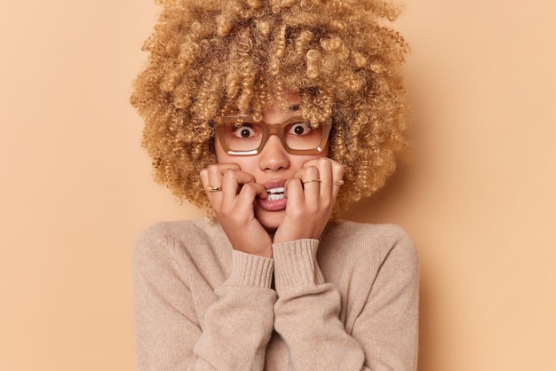 Photo young scared young woman bites finger nails looks anxious and worried wears transparent glaasses and jumper isolated over beige background suffers from phobia negative human emotions concept