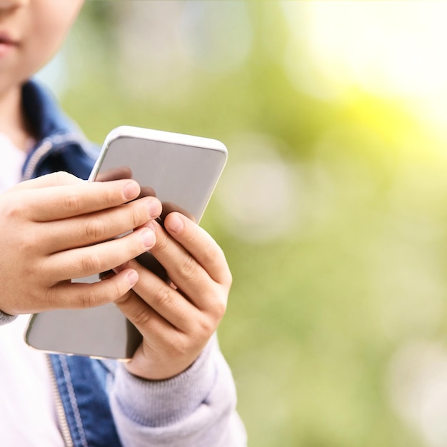 Young school boy texting outdoor Male person using smartphone Little hands with cell phone Green bokeh street background